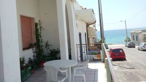 Un balcon avec une table et des chaises, donnant sur une rue bordée de voitures garées et offrant une vue sur la mer.