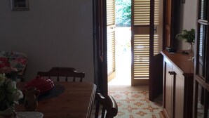 Un coin repas avec une table en bois, des chaises et une petite table sur laquelle se trouvent une carafe et des fleurs. Une porte donne accès à un balcon d’où l’on peut admirer la verdure.