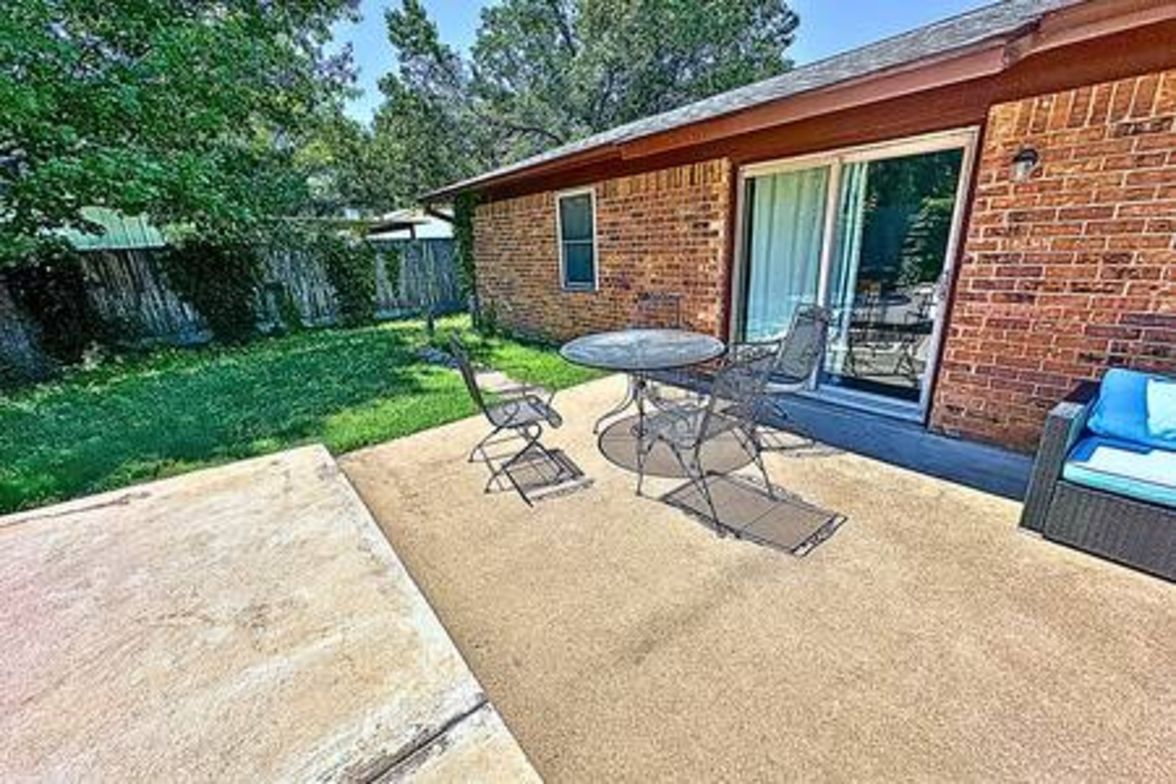 A patio with a table and chairs, a brick building with a sliding glass door, and a grassy yard.