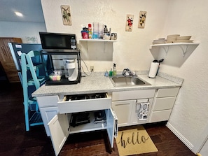 A compact kitchen with a microwave, coffee maker, and a sink. There are wall-mounted shelves with dishes and a 'hello' mat on the floor.
