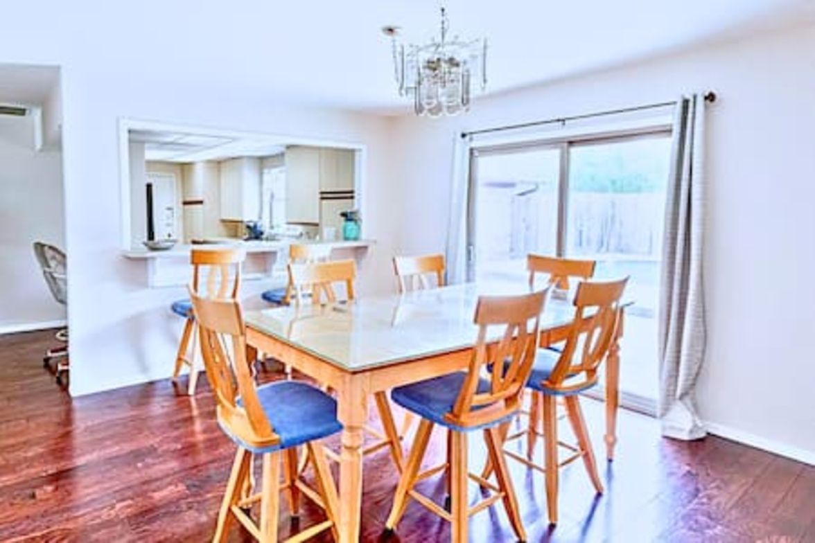 A dining area with a rectangular table surrounded by wooden chairs with blue cushions, a chandelier, and a large window with curtains.