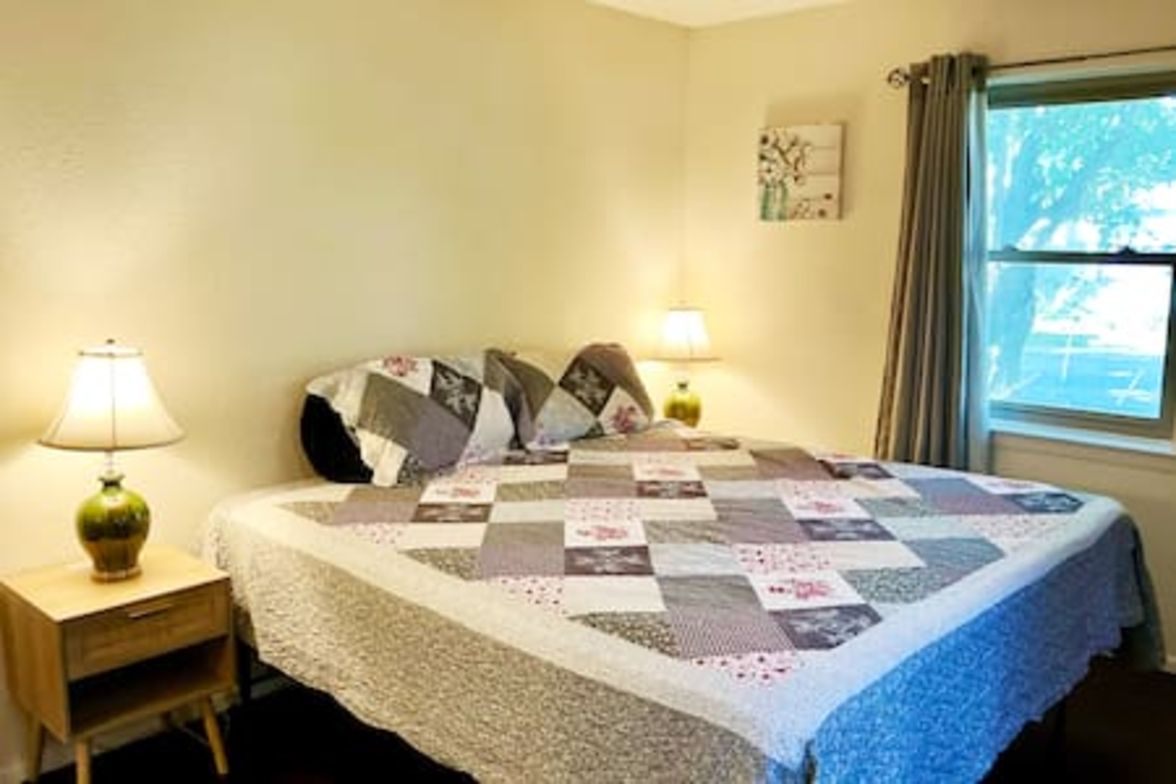 A bedroom with a bed, two bedside lamps, a window with curtains, and a small bedside table.