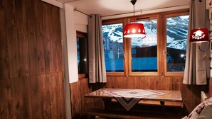 Una habitación acogedora con paredes y mesa de madera, y un banco. Hay una ventana con cortinas, una lámpara colgante y vistas a una montaña nevada.