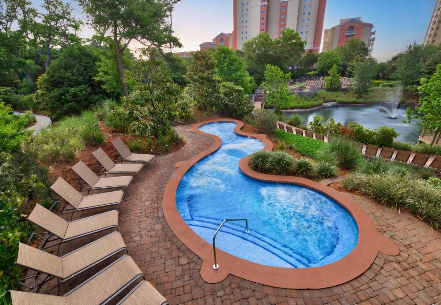 A swimming pool with a winding design, surrounded by lush greenery and a brick pathway.