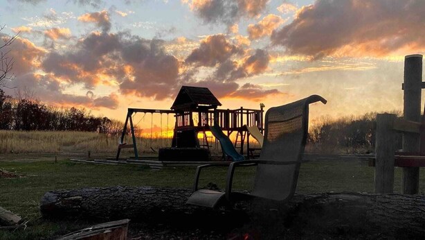 A fire pit with rocks, a swing set, and a bench in a grassy area with a sunset in the background. - The Hruby Doobie Doo Ranch (Crown Point)
