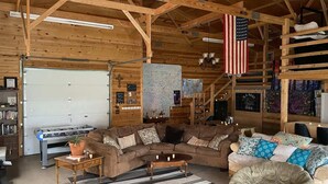 A spacious living room with a large couch, a coffee table, and a chair. There is a flag hanging from the ceiling, a bookshelf, and a map on the wall. - The Hruby Doobie Doo Ranch (Crown Point)