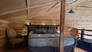 A cozy bedroom with a wooden ceiling, a bed with patterned bedding, a chair, a bedside table with a lamp, and a heater. - The Hruby Doobie Doo Ranch (Crown Point)