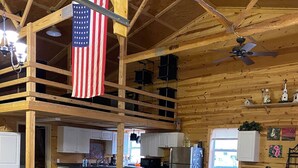 A spacious loft-style living area with a high ceiling, wooden beams, a large American flag, a fireplace, and a cozy seating arrangement. - The Hruby Doobie Doo Ranch (Crown Point)
