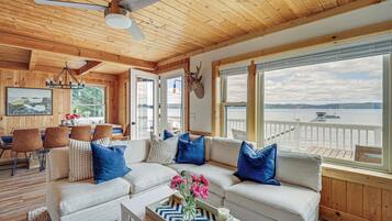 L’intérieur chaleureux d’un chalet avec un plafond en bois, une grande fenêtre donnant sur un lac, un canapé avec des coussins bleus et blancs, et un coin repas avec des chaises et une table.