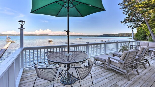 A wooden deck with a round table and chairs, a green umbrella, and a view of the water. - Updated Glen Arbor Cottage on Lake: Dock & Mooring (Glen Arbor)