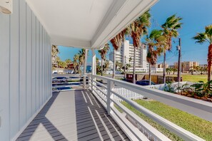 A covered outdoor area with a wooden deck, white railings, and a view of palm trees and buildings in the background.