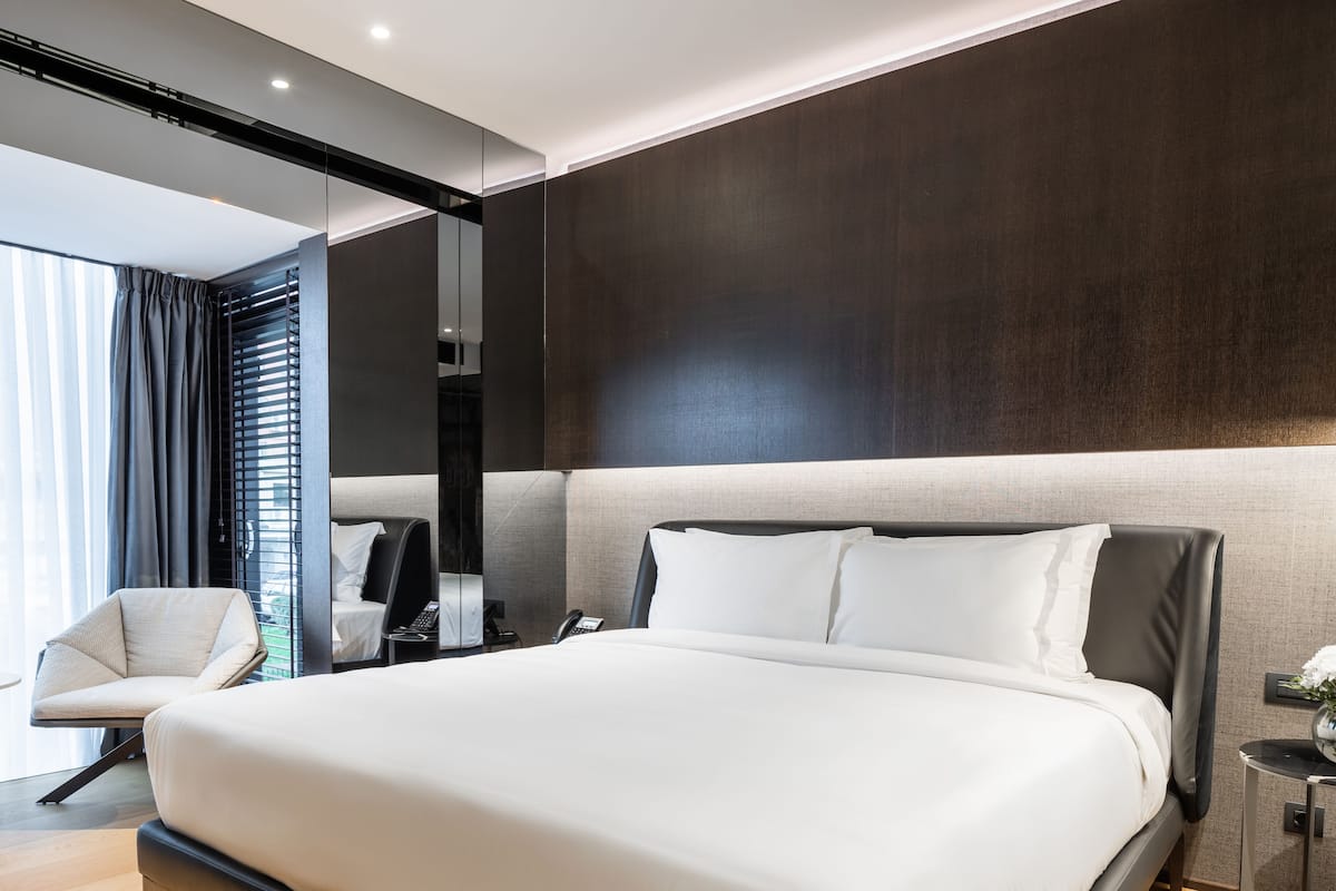 A modern hotel room with a large bed, a chair, a bedside table, and a mirror.