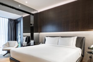 Quarto de hotel moderno com uma cama grande, uma cadeira, uma mesinha de cabeceira e um espelho.