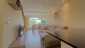 Ampla sala de estar com sofá, mesa de centro e TV sobre suporte de madeira. Há uma área de jantar com cadeiras e mesa, e uma grande janela com vista para o exterior.