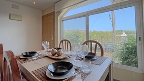 Mesa posta com pratos, tigelas e copos, com vista para áreas verdes e edifícios por uma grande janela.