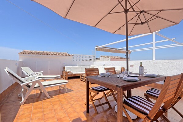 A rooftop terrace with a wooden table and chairs, a large umbrella, and a white sofa. - 053 - Altomar II 003 - comfortHOLIDAYS (Santa Pola)