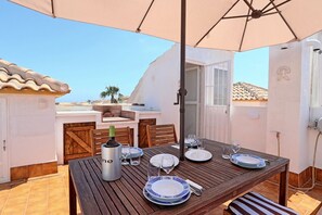 A dining area with a wooden table set for four, a wine bottle, and a large umbrella providing shade. - 053 - Altomar II 003 - comfortHOLIDAYS (Santa Pola)