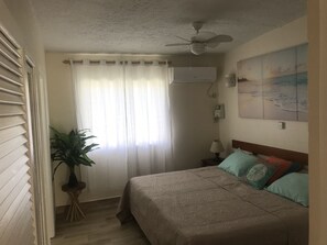 A bedroom with a bed, a ceiling fan, a nightstand with a lamp, a potted plant, and a painting on the wall.