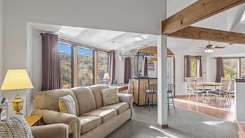 A spacious living area with a sofa, dining table, and chairs, large windows with a view, and a ceiling fan.