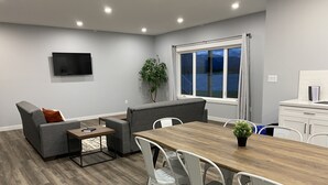 Een moderne woonkamer met een bank, een eettafel en stoelen. Er is een tv aan de muur bevestigd en een raam met uitzicht op het omliggende landschap.