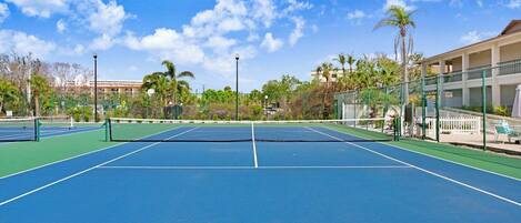 An outdoor tennis court with a net, surrounded by a fence and palm trees.