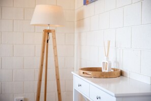 Un mur en briques blanches, un lampadaire blanc, une commode blanche avec des tiroirs, et un plateau en bois avec un porte-bougie blanc et un diffuseur de parfum à bâtonnets.