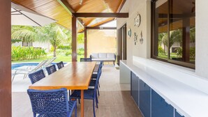 A covered outdoor dining area with a wooden table and blue chairs, a white bench, and a view of a swimming pool and greenery. - OKA BEACH RESIDENCE Porto de Galinhas (Ipojuca)