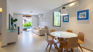 A modern living room with a white sofa, a glass coffee table, and wooden chairs. There is a dining area with a white table and chairs. The room has large windows and a view of greenery outside. - OKA BEACH RESIDENCE Porto de Galinhas (Ipojuca)