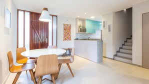 A modern kitchen with a central island, wooden chairs around a round table, and a staircase to the right. - OKA BEACH RESIDENCE Porto de Galinhas (Ipojuca)