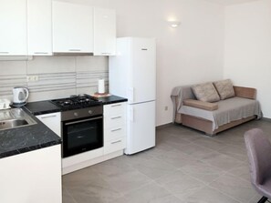 A compact kitchen with white cabinets, a stove, oven, and a refrigerator. There's a sofa with a blanket and pillows in the adjacent living area.