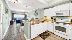 A kitchen with white cabinets, a microwave, and an oven. There is a clock on the wall and a window overlooking a balcony. - Crystal Tower 1603, Gulf views/ 3 Pools/King Bed (Gulf Shores)