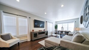 Una sala de estar moderna con un televisor de pantalla plana, una zona de comedor con mesa y sillas, y un ventanal con persianas.