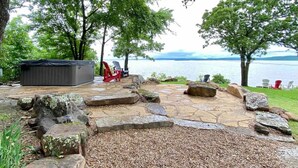 A stone patio with a hot tub, red Adirondack chairs, and a view of a lake.