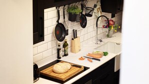 Cozinha com azulejos brancos, panelas penduradas, tábua de corte com pão e diversos utensílios de cozinha.