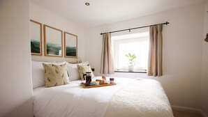 Quarto com uma cama grande, mesa de cabeceira com cafeteira e doces, e janela com cortinas.