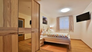 Een slaapkamer met een houten garderobe, een bed met een stippenbedekking, een aan de muur bevestigde televisie en een raam met gordijnen.