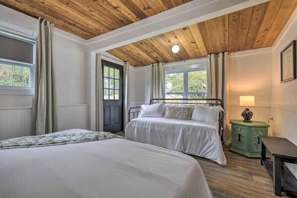 A bedroom with two beds, wooden ceiling, a green nightstand, and a window with curtains. - Sip, Stay & Gather! Vintage Hill Country Bungalow (Fredericksburg)