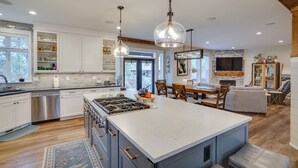 A modern kitchen with white cabinets, stainless steel appliances, and a large island. There is a dining area with a wooden table and chairs, and a fireplace.