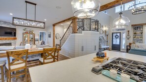 A spacious kitchen with a large island, wooden chairs, and a dining table. There is a staircase leading to an upper level, and the kitchen features modern lighting fixtures.