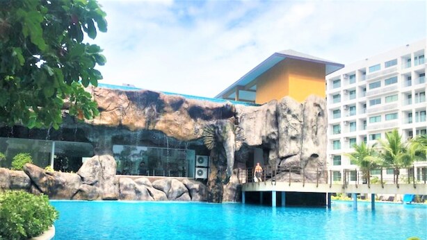 A swimming pool with a rocky waterfall feature, a glass bridge, and a modern building in the background. - Laguna Beach 3 The Maldives - top floor studio (Pattaya)
