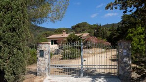 A gated entrance to a property with a stone wall and metal gate, surrounded by trees and a hill in the background. - Independent Villa l'Aia in a panoramic and quiet position on the Gulf of Lacona (Capoliveri)