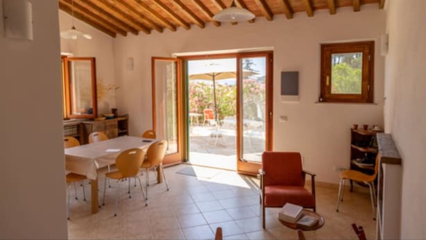 A spacious room with a wooden ceiling, a dining area with a table and chairs, a red armchair, and a sliding glass door leading to an outdoor patio with umbrellas. - Independent Villa l'Aia in a panoramic and quiet position on the Gulf of Lacona (Capoliveri)