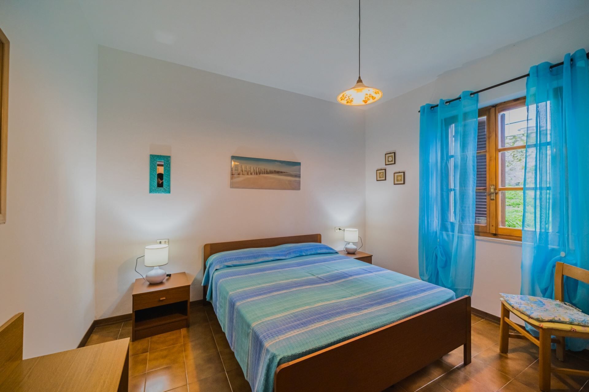 A bedroom with a bed, bedside tables, a lamp, a painting, and a window with curtains.