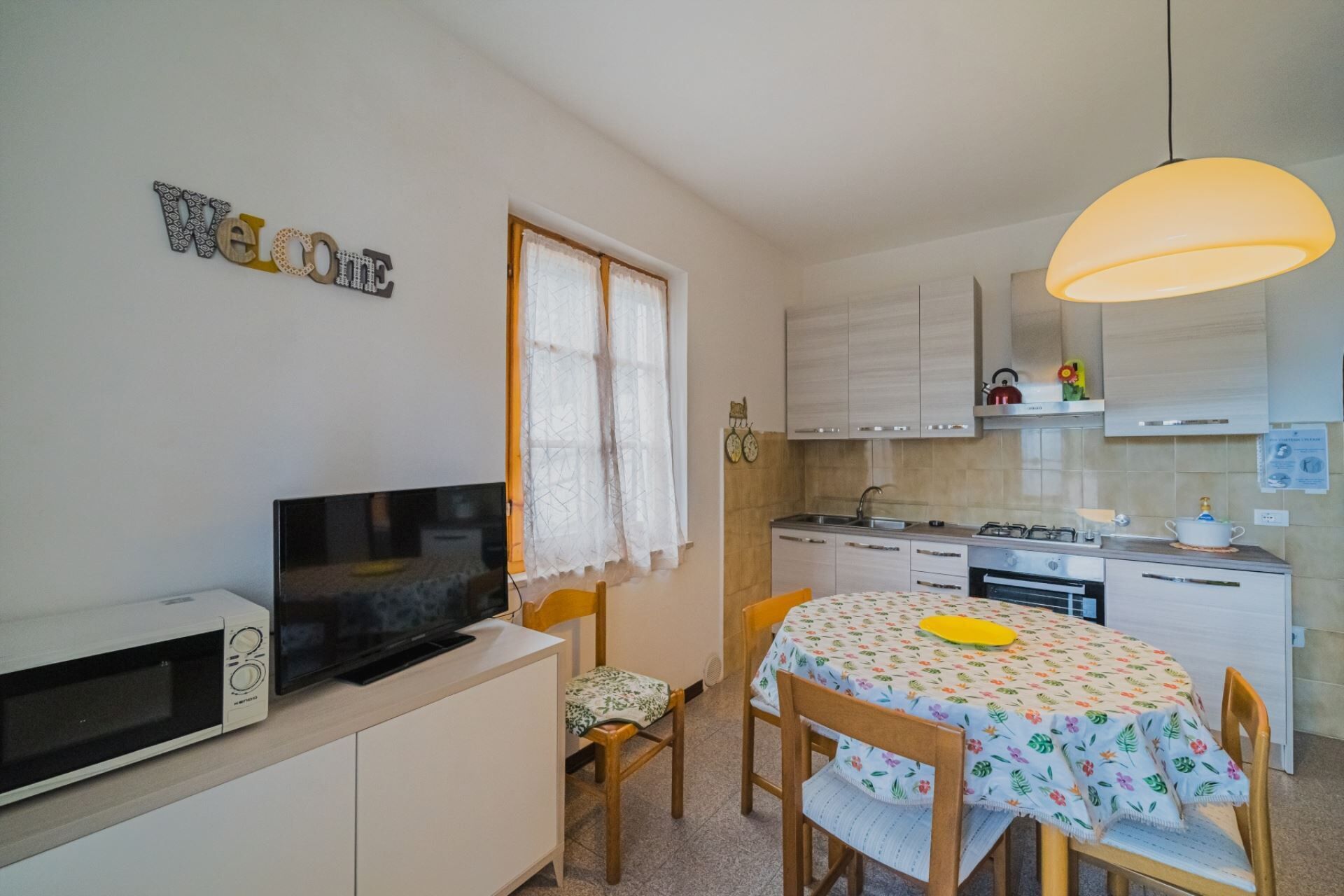 A compact kitchen with a microwave, stove, and sink. A TV is mounted on the wall, and a 'Welcome' sign is visible.