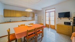 A compact kitchen with wooden cabinets, a dining table with orange chairs, a built-in microwave, and a television on a shelf. - APPARTAMENTI SANT'ANDREA - CASA GRANDE ALLE PIANE (Marciana)