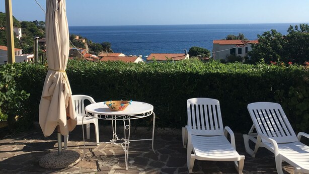 A patio with stone flooring, white chairs, a table with a bowl, and an umbrella. - APPARTAMENTI SANT'ANDREA - CASA GRANDE ALLE PIANE (Marciana)