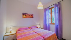 Ein Doppelbett mit gestreifter Bettwäsche, ein Nachttisch mit Lampe, ein Fenster mit violetten Vorhängen und ein Sonnenuntergangsbild an der Wand.