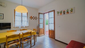 Ein Essbereich mit einem gelben Tisch und Stühlen, einem Fernseher auf einem Standfuß und einem großen Fenster mit einer gelben Lampe darüber.