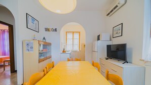 Ein helles Zimmer mit einem gelben Tisch, Stühlen, einem Fernseher und einem Kühlschrank.