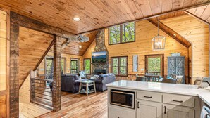 A log cabin interior with a stone fireplace, wooden floors, a dining area, and a kitchen with a large island.